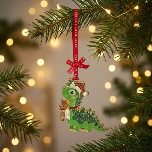 A whimsical Tipperary Crystal Sparkle dinosaur Christmas tree ornament hanging from a branch. The gold-trimmed green metal dinosaur is shaped like a brontosaurus, wearing a red and white enamel Santa hat with holly leaves, a red and white striped scarf, and featuring a row of shimmering green crystal rhinestones embedded along its back. It hangs from a red fabric ribbon printed with "Tipperary Crystal" text, surrounded by blurry golden background holiday lights.