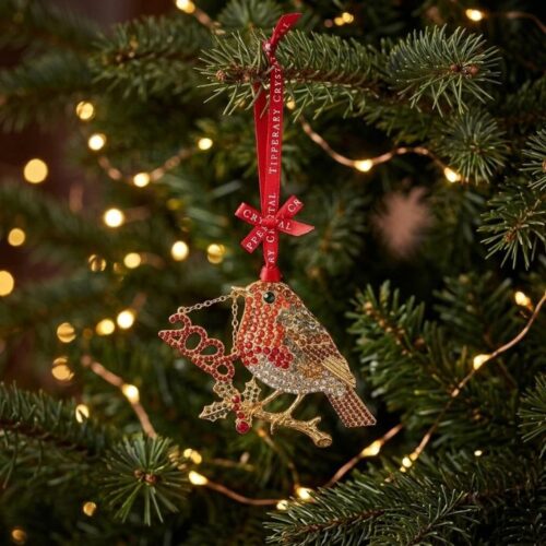 A sparkling Tipperary Crystal Christmas robin ornament hanging on a pine tree branch. The gold-toned robin is covered in brilliant red and silver crystal gemstones, perched on a branch with green holly leaves and red berries. It holds a delicate chain with a red crystal "2026" dated charm. The ornament hangs from a red silk ribbon with the words "Tipperary Crystal" printed on it, surrounded by warm, glowing Christmas tree lights in the background.