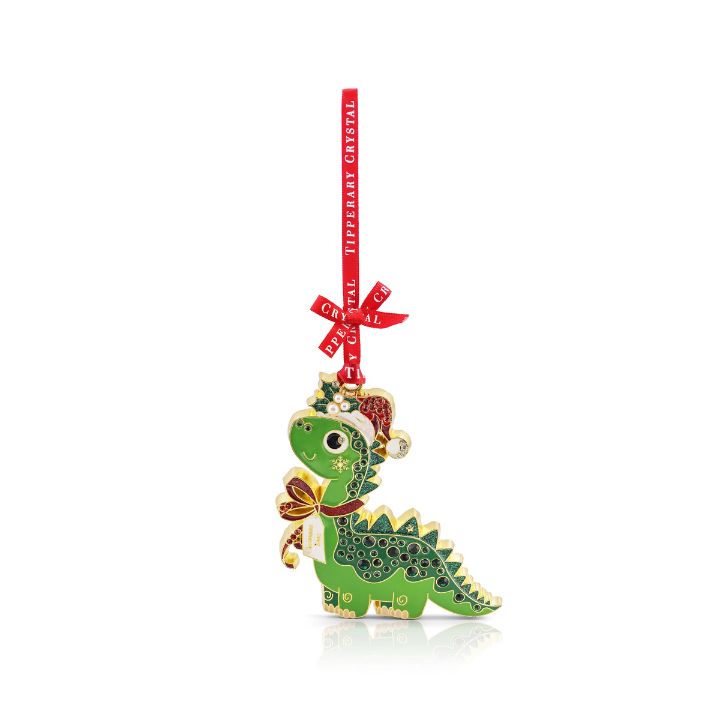 A whimsical Tipperary Crystal Sparkle dinosaur Christmas tree ornament hanging from a branch. The gold-trimmed green metal dinosaur is shaped like a brontosaurus, wearing a red and white enamel Santa hat with holly leaves, a red and white striped scarf, and featuring a row of shimmering green crystal rhinestones embedded along its back. It hangs from a red fabric ribbon printed with "Tipperary Crystal" text, surrounded by blurry golden background holiday lights.