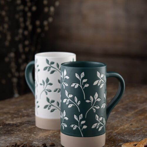 • • Two tall Belleek Living Flora fine china mugs with green and white botanical leaf patterns standing on a rustic wooden table.