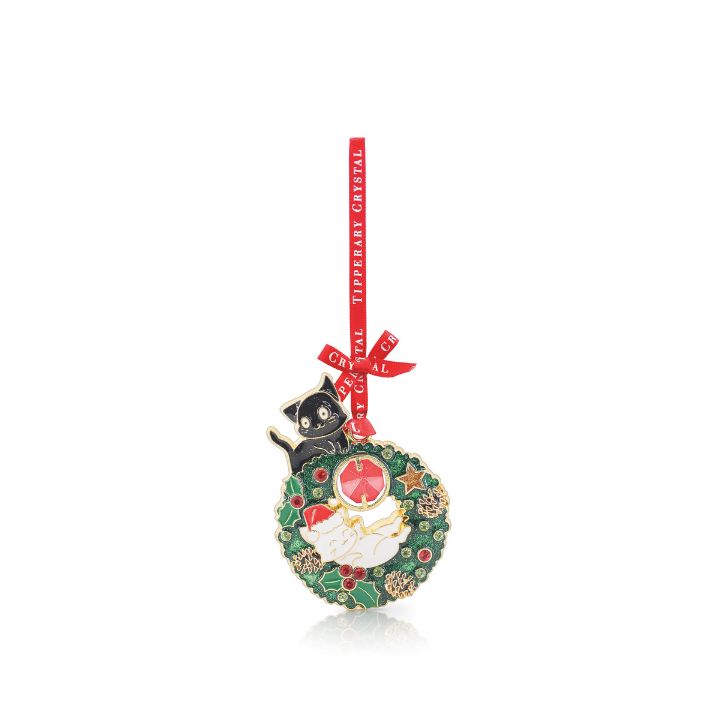 e holiday ornament features an intricate green enamel Christmas wreath accented with golden stars and pinecones, a sleeping white cat wearing a Santa hat nestled in the center, and a wide-eyed black cat perched on top. A vibrant red ribbon printed with "Tipperary Crystal" is tied in a neat bow at the top for hanging.