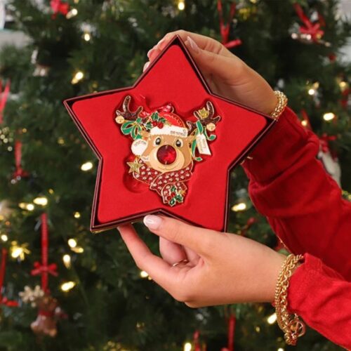 A close-up view of hands holding an open star-shaped red gift box displaying a Tipperary Crystal Gem Reindeer Christmas tree ornament featuring a sparkling red nose, a crystal-embellished scarf, and a "Merry Christmas" sign in front of a blurred background tree with warm lights.