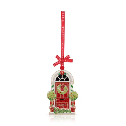 A festive red front door Christmas ornament by Tipperary Crystal decorated with a green holiday wreath, two flanking topiary potted trees, and the text "Home for Christmas" written at the base, hanging from a branded red ribbon.